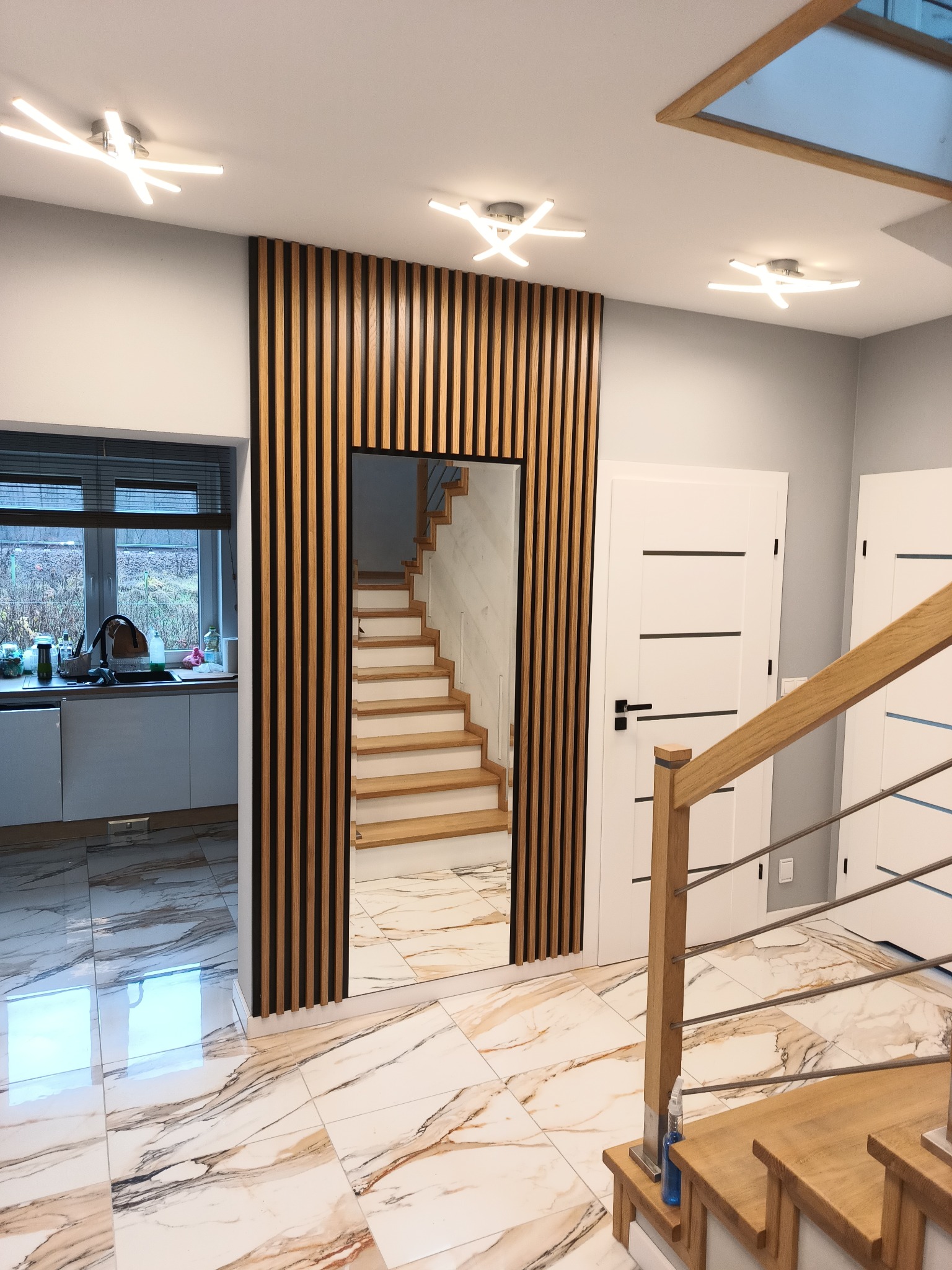Nowoczesny przedpokój z lustrem otoczonym drewnianymi lamelami, widok na schody i fragment zabudowy kuchennej z oknem.