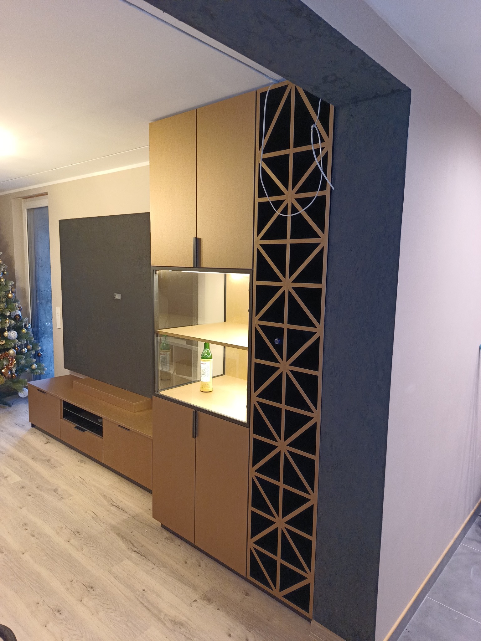 Nowoczesna zabudowa meblowa w salonie z szafkami z forniru w odcieniach brązu, szarym panelem TV oraz dekoracyjnym ażurem z trójkątnych motywów. W tle widoczna choinka.