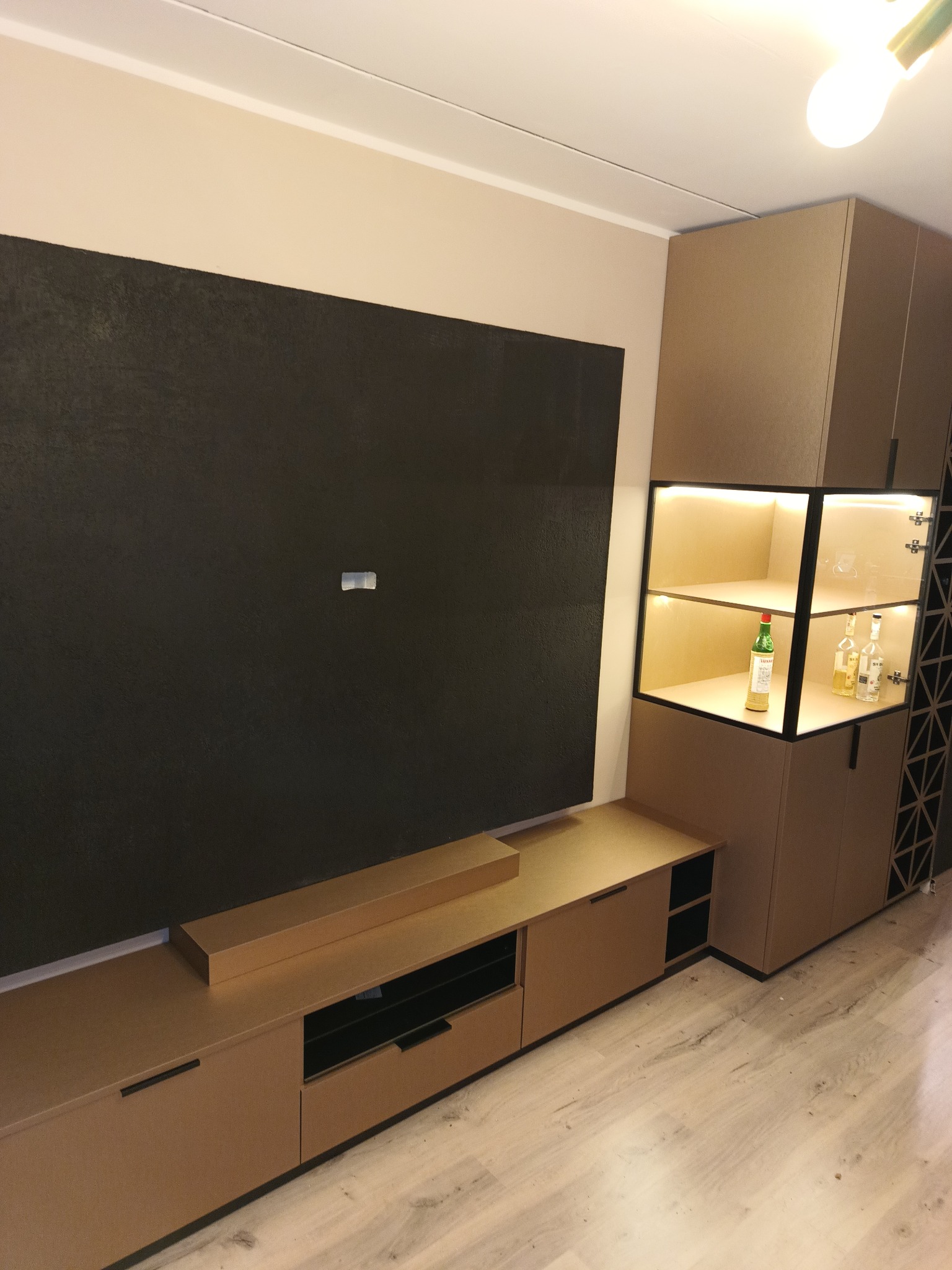 Nowoczesna zabudowa meblowa w salonie z ciemną ścianą, oświetloną witryną z alkoholem i szafkami w odcieniach beżu i brązu.
