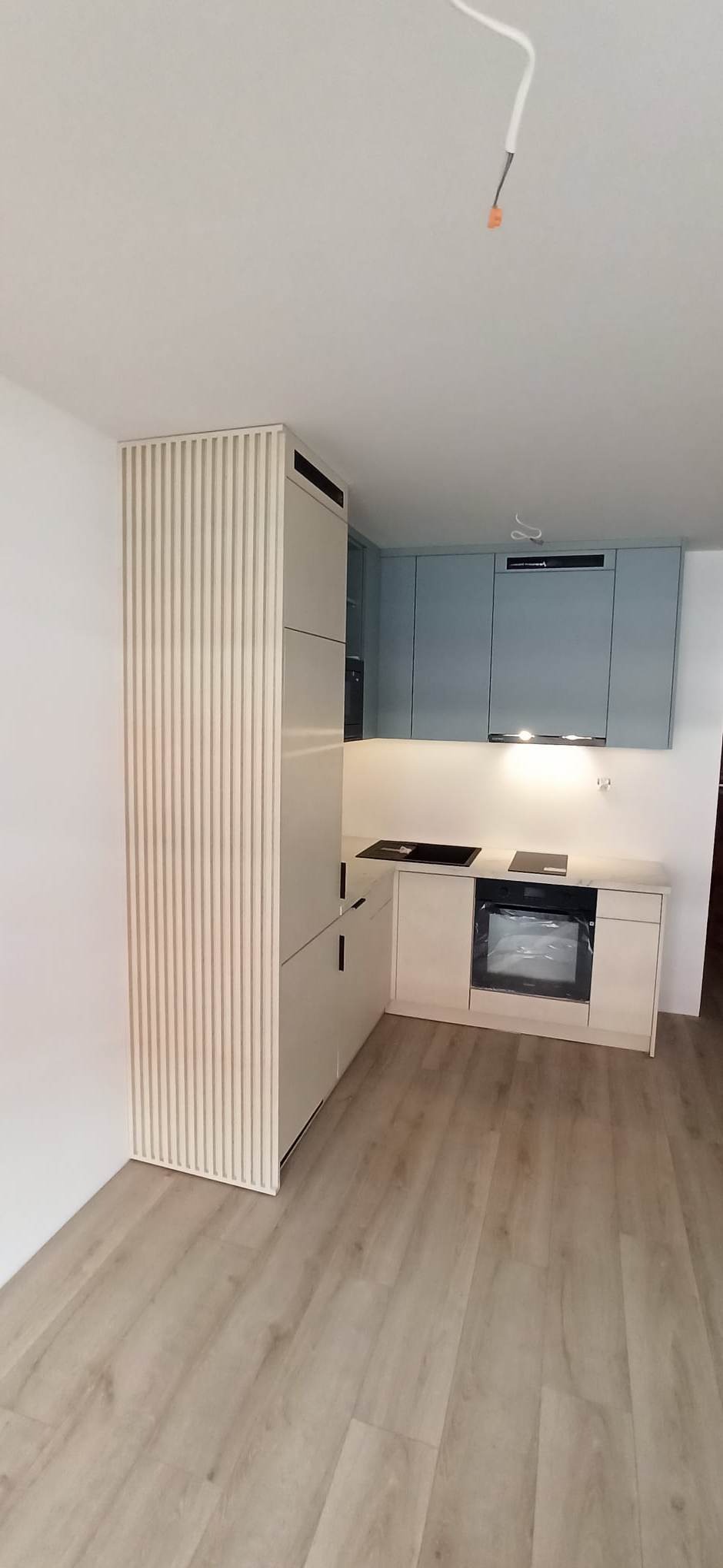 Jasna kuchnia w kształcie litery L z frontami w kolorze beżowym i błękitnym, z pionowymi żłobieniami na wysokiej szafce, płyta indukcyjna i piekarnik w zabudowie, oświetlenie podszafkowe.