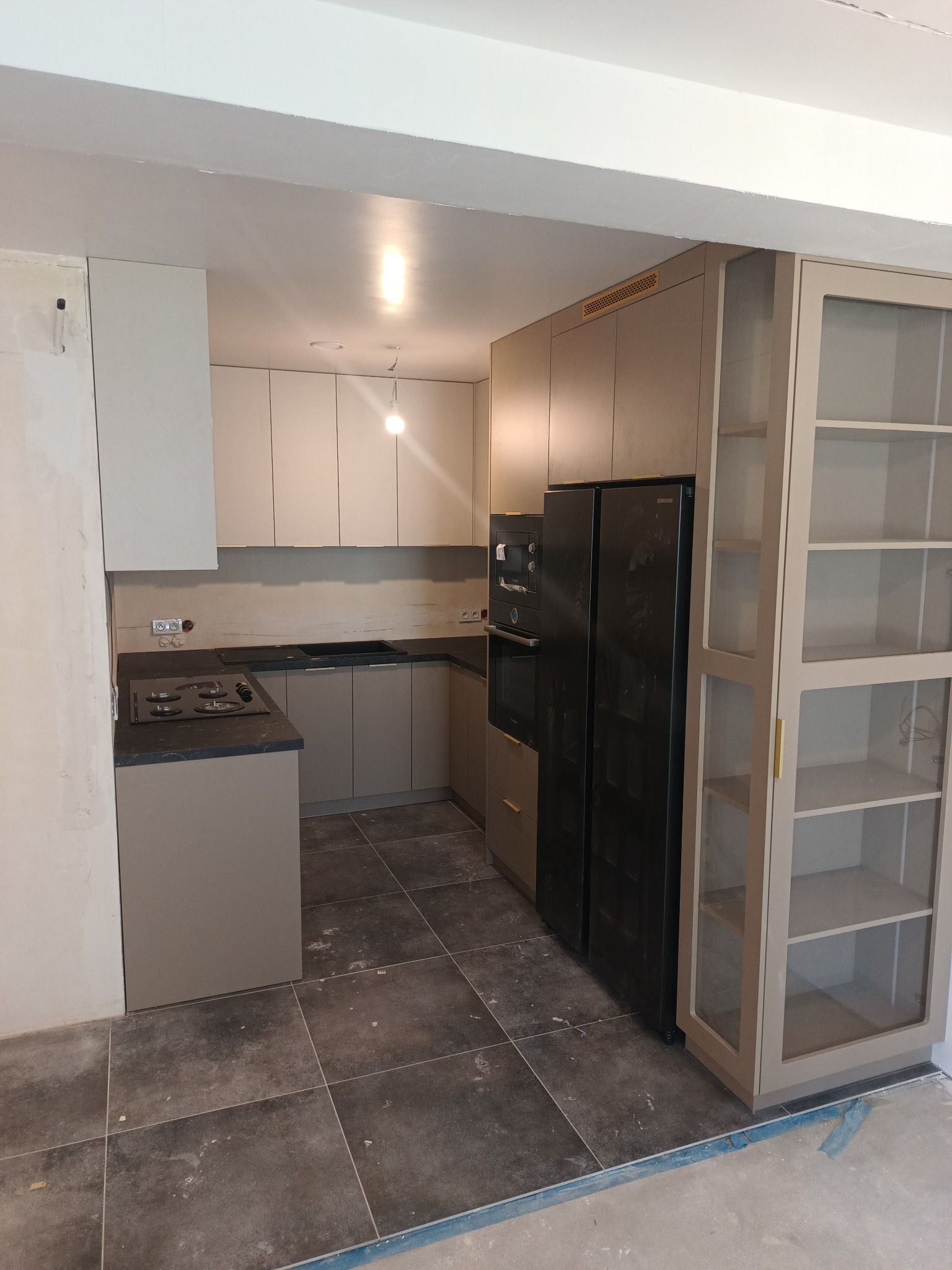 Wnętrze kuchni w trakcie remontu, z zamontowanymi beżowymi szafkami kuchennymi, czarnym sprzętem AGD i szarą podłogą z płytek, widoczne niedokończone ściany.