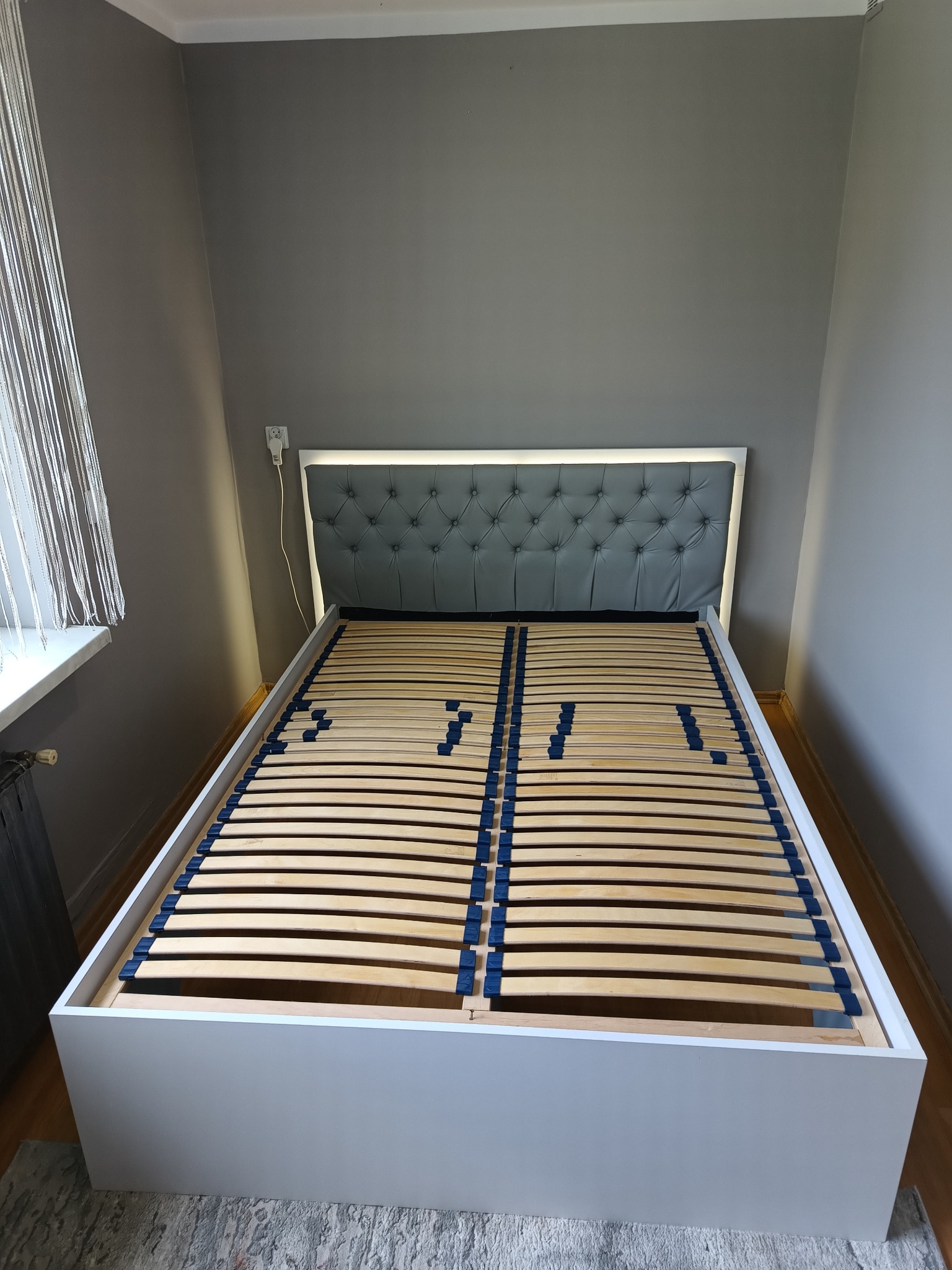 Białe łóżko z szarym, pikowanym zagłówkiem z podświetleniem LED, widoczny stelaż z drewnianych listew.