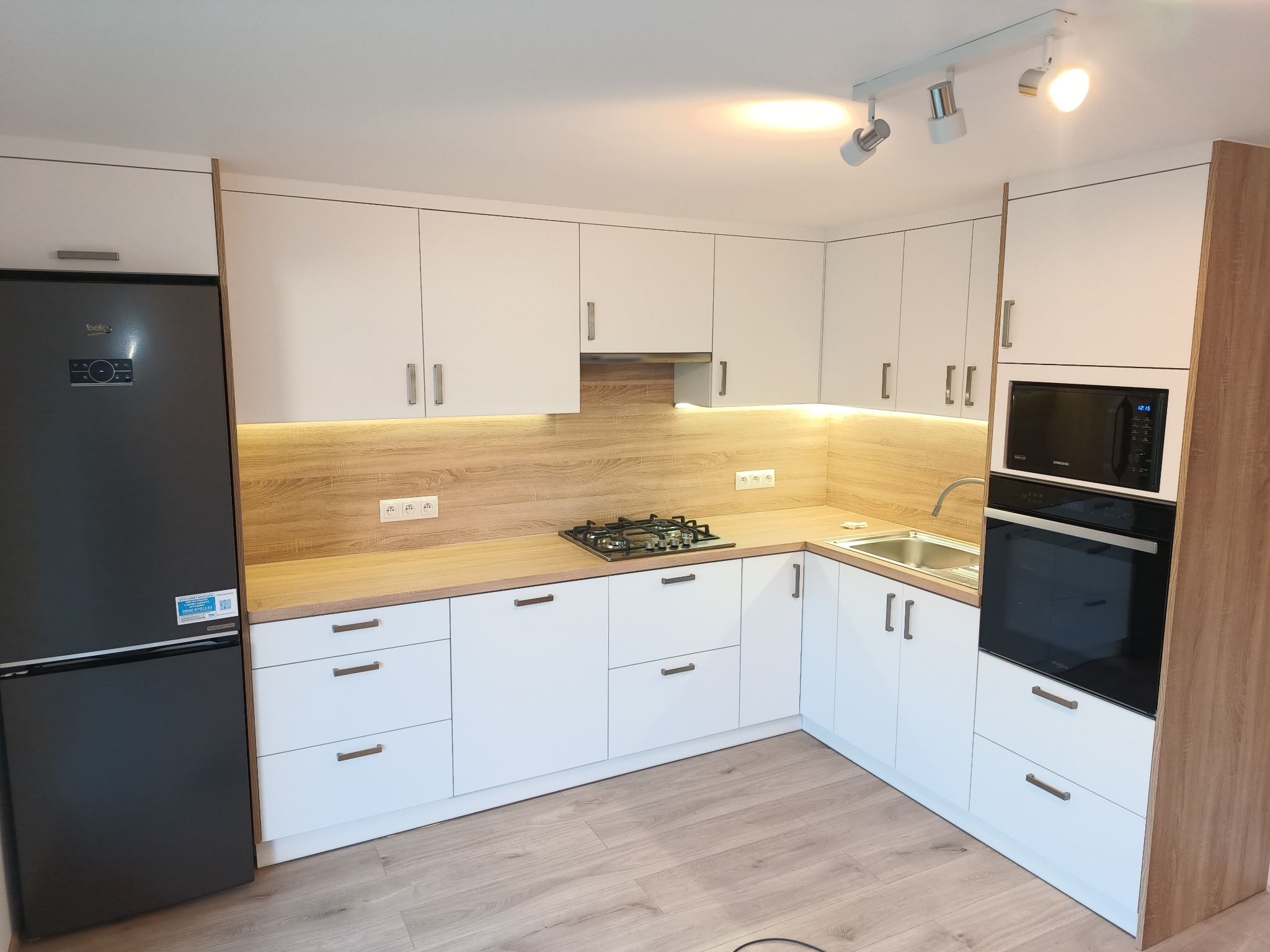 Nowoczesna kuchnia z białymi frontami i drewnianym blatem, wyposażona w czarną lodówkę, płytę gazową i piekarnik z mikrofalówką w zabudowie, oświetlona punktowymi lampami sufitowymi.