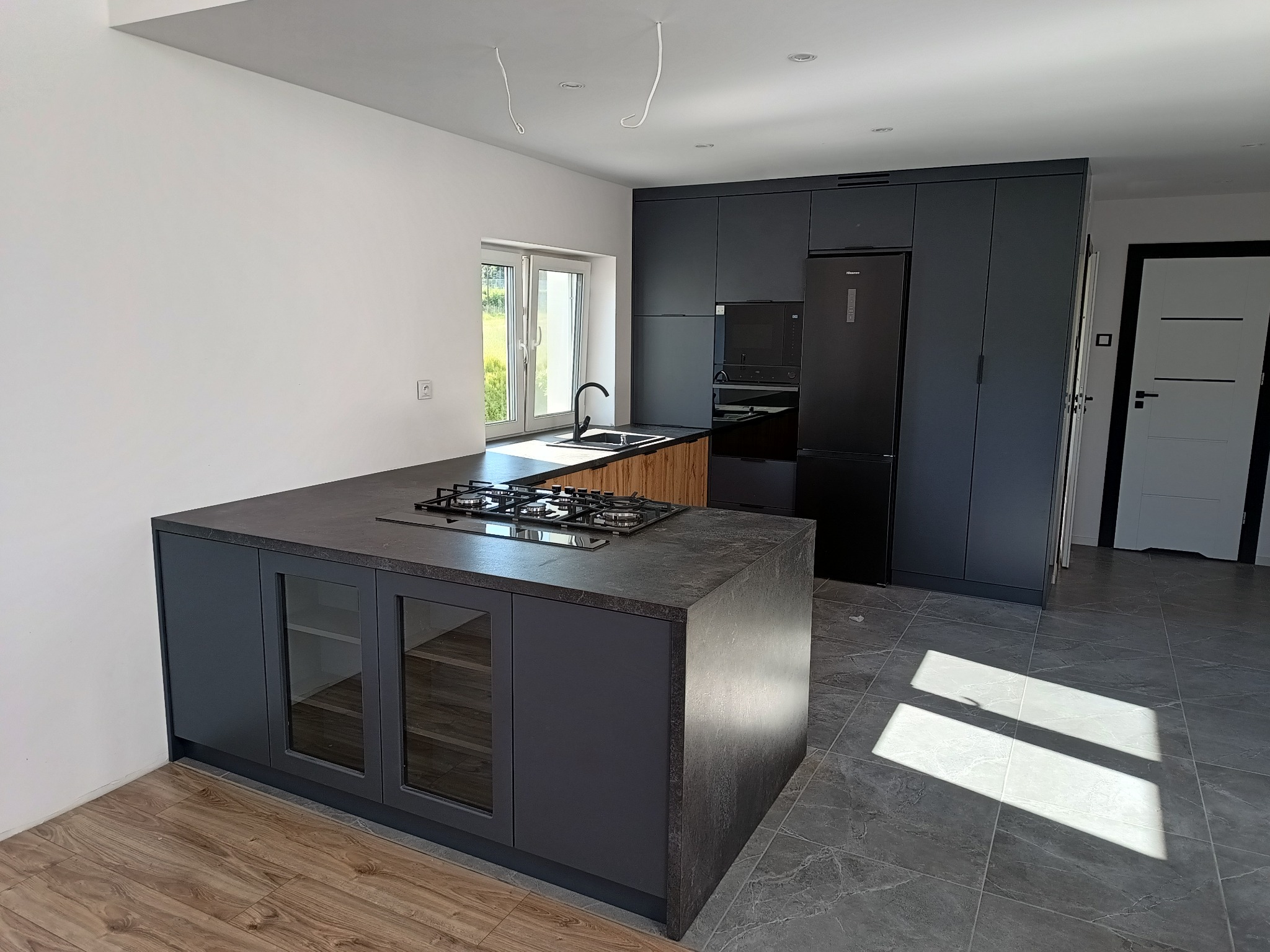 Nowoczesna kuchnia z szarymi szafkami, wyspą z płytą gazową i przeszklonymi frontami, czarnym zlewem przy oknie i czarną lodówką Hisense.