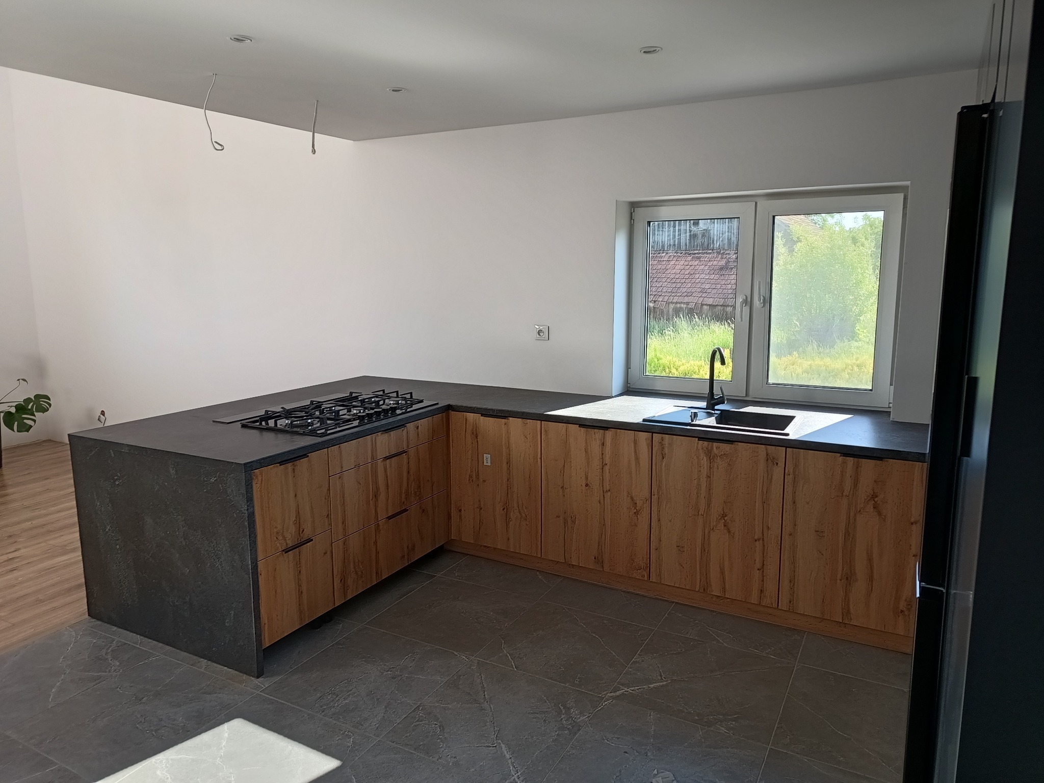 Nowoczesna kuchnia z drewnianymi frontami szafek i ciemnym blatem, płyta gazowa wbudowana w blat, widok na okno z zielenią na zewnątrz, szara podłoga z płytek, brakujące lampy wiszące.