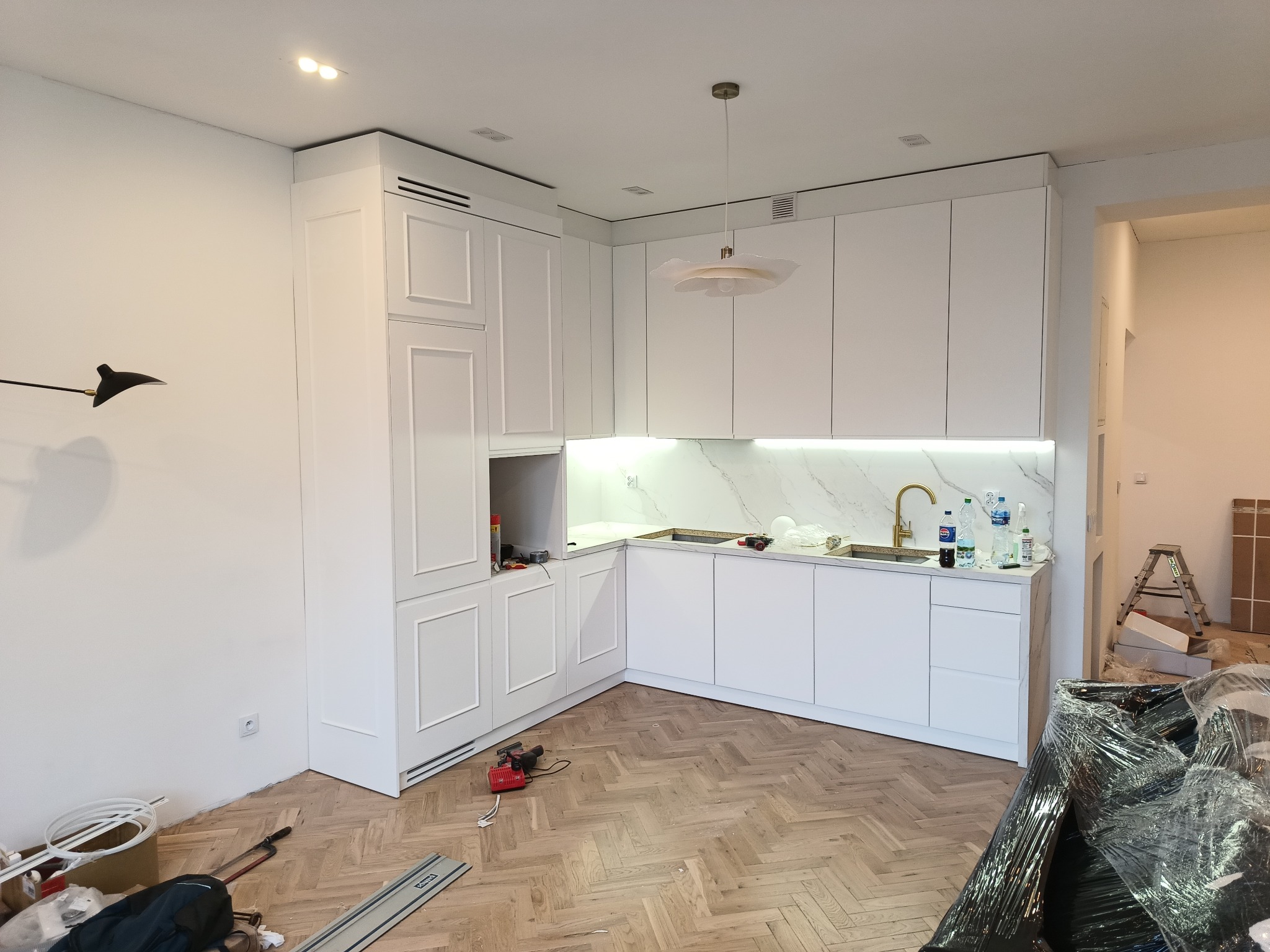 Wnętrze kuchni w trakcie montażu: białe fronty szafek z frezowanymi detalami, marmurowy backsplash, złota bateria, narzędzia i materiały budowlane na parkiecie ułożonym w jodełkę.