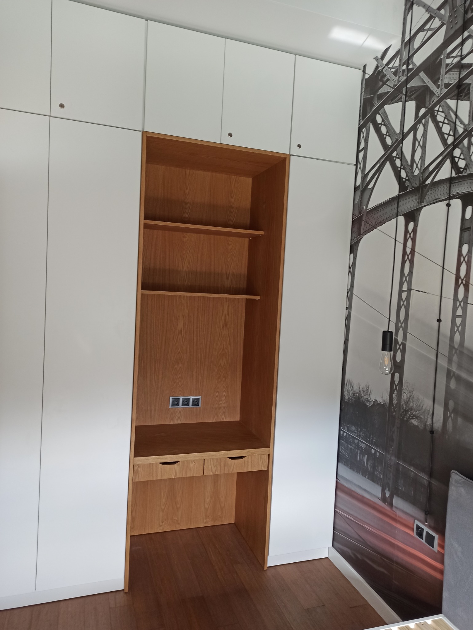 Biała zabudowa meblowa z drewnianym regałem z półkami i biurkiem, widoczne gniazdka elektryczne, na ścianie fototapeta z motywem mostu.