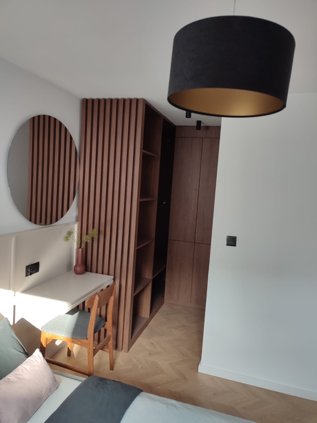 Nowoczesna szafa wnękowa z drewnianymi lamelami i otwartymi półkami, zintegrowana z biurkiem i okrągłym lustrem w minimalistycznym wnętrzu sypialni, oświetlona wiszącą lampą z czarnym abażurem...
