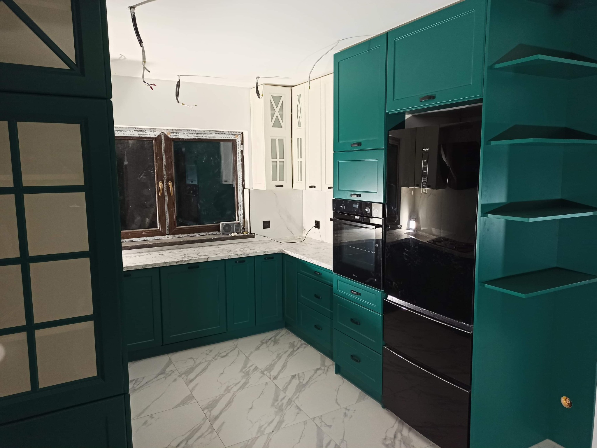 Szmaragdowa zabudowa kuchenna z czarnym piekarnikiem i marmurowym blatem w Woli Radziszowskiej, widoczne niedokończone podłączenia elektryczne sugerujące montaż.