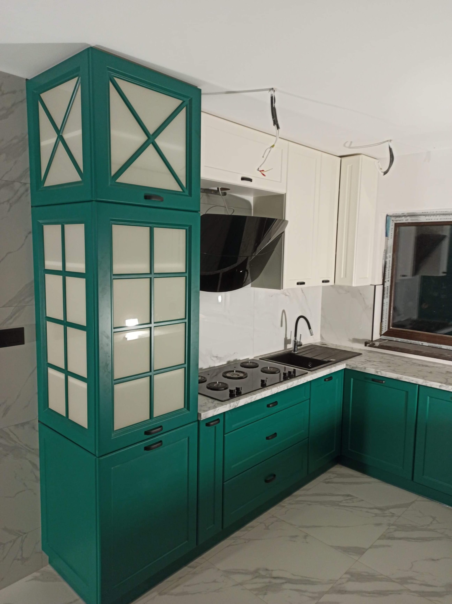 Nowoczesna kuchnia w kolorze butelkowej zieleni i bieli, z marmurowym blatem i płytkami, widoczne niedokończone podłączenia elektryczne.