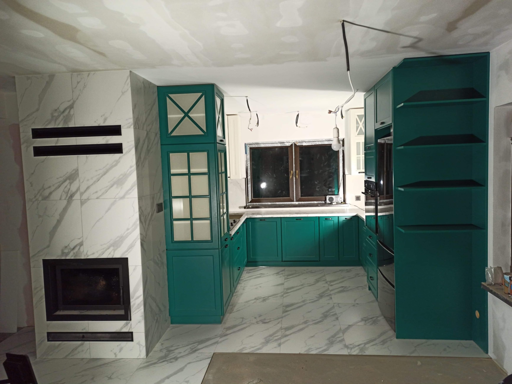 Kuchnia w trakcie montażu: zielone szafki z przeszklonymi witrynami, marmurowe płytki na podłodze i obudowie kominka, widoczne niedokończone instalacje elektryczne.