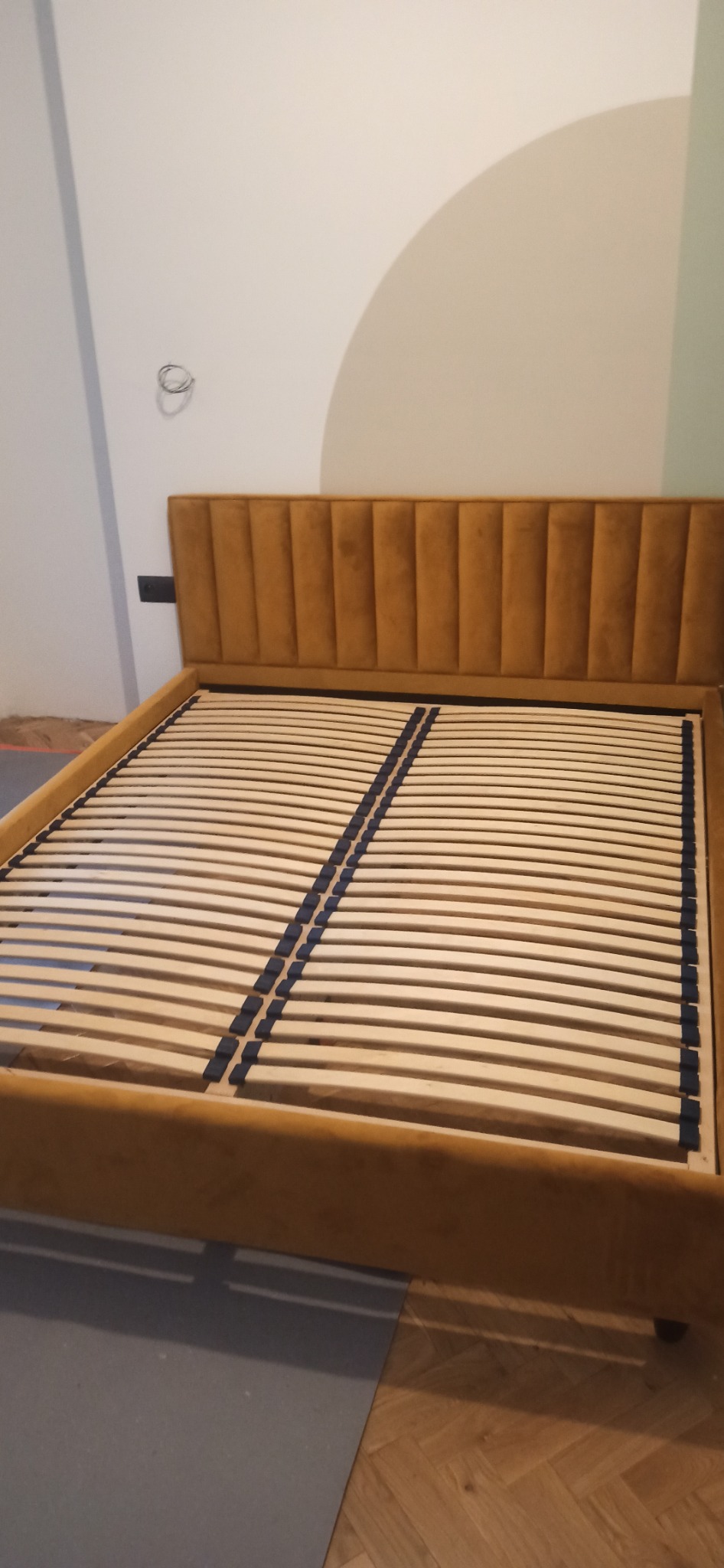 Rama łóżka z tapicerowanym, musztardowym zagłówkiem w pionowe pasy i drewnianym stelażem listwowym, widok z przodu na tle ściany w trakcie malowania.