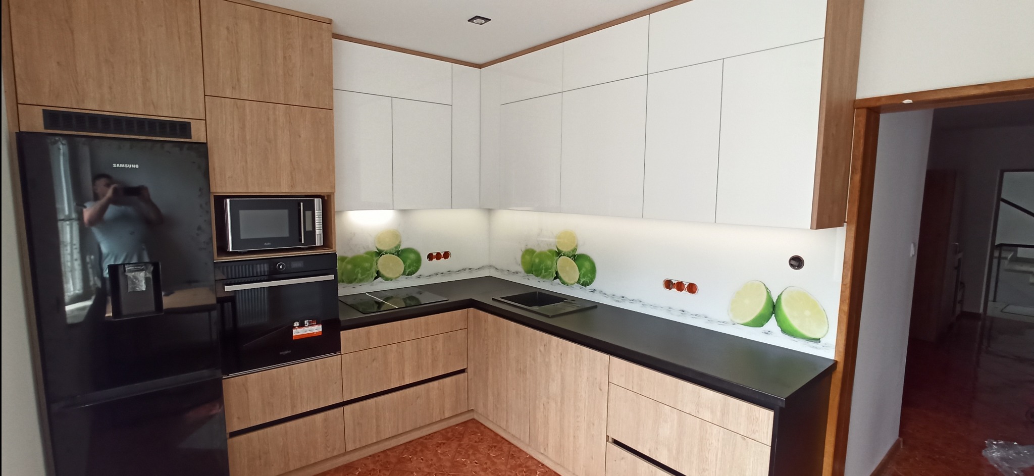 Nowoczesna kuchnia z frontami w kolorze jasnego drewna i białymi szafkami górnymi, czarnym blatem i panelem szklanym z motywem limonek. W zabudowie czarna lodówka Samsung z odbijającym się fotografem.