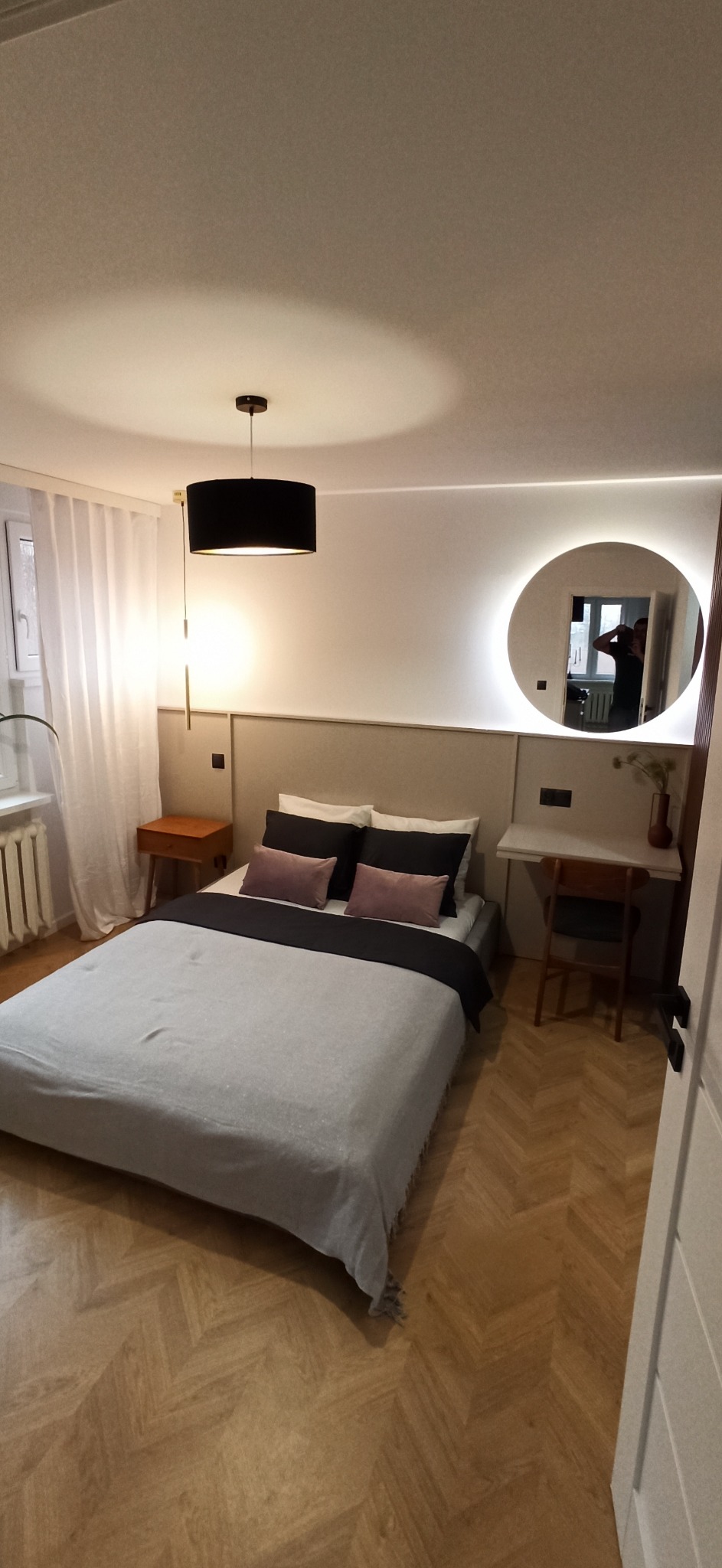 Nowoczesna sypialnia z łóżkiem, drewnianą podłogą ułożoną w jodełkę, okrągłym lustrem z podświetleniem LED i biurkiem z krzesłem.