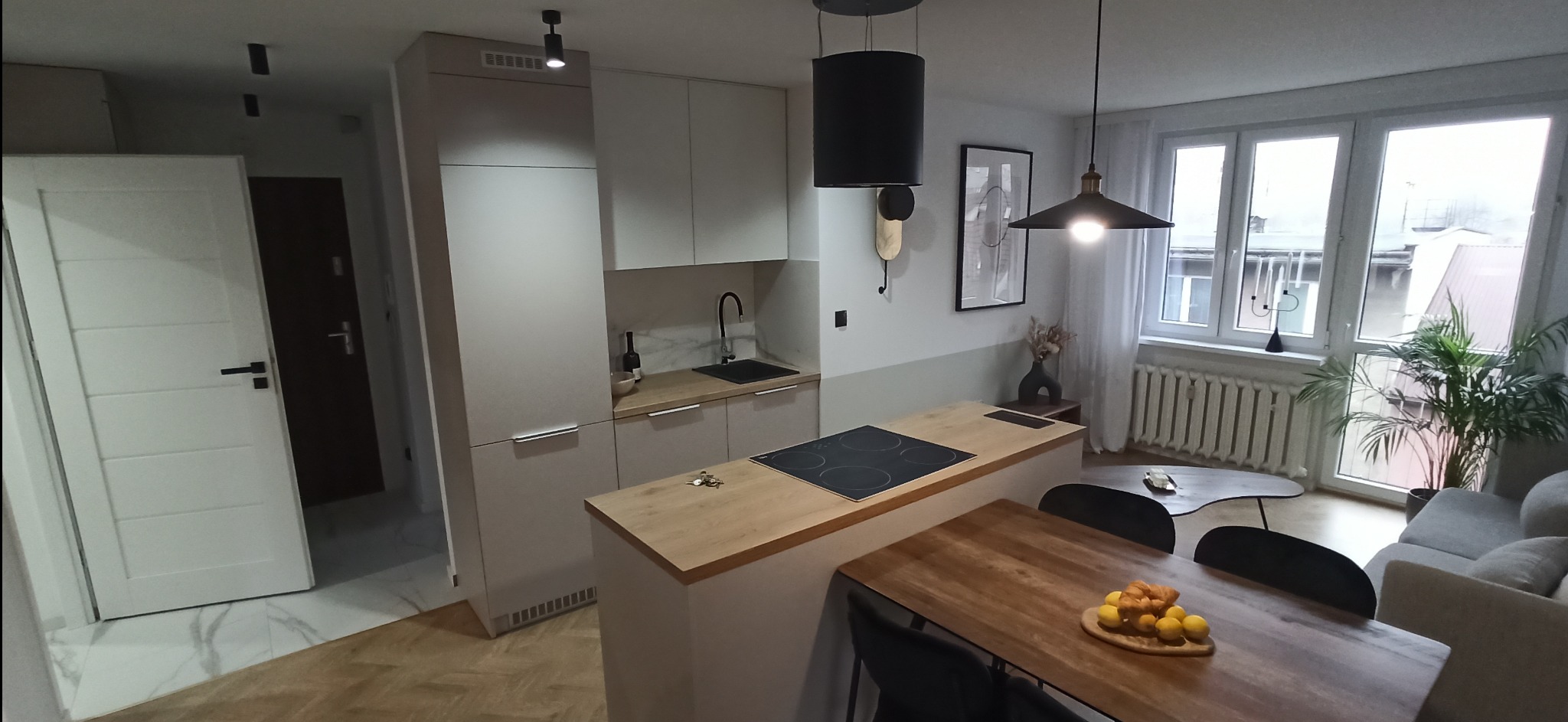 Nowoczesna kuchnia z jasnymi szafkami, drewnianym blatem i wyspą z płytą indukcyjną, połączona z jadalnią z drewnianym stołem i owocami na blacie, widok na salon z szarą sofą i roślinami w tle.