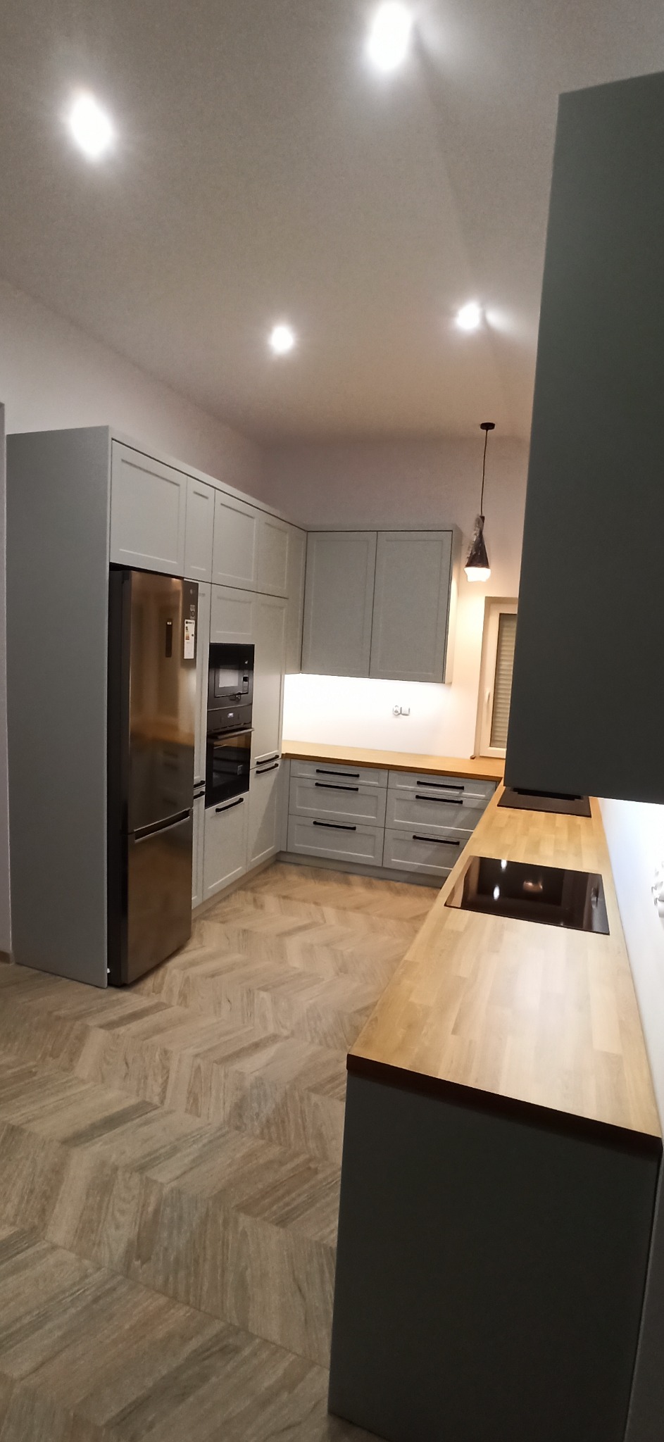 Kompletna kuchnia w odcieniach szarości z drewnianymi blatami i parkietem ułożonym w jodełkę; widoczna lodówka, piekarnik i płyta indukcyjna.