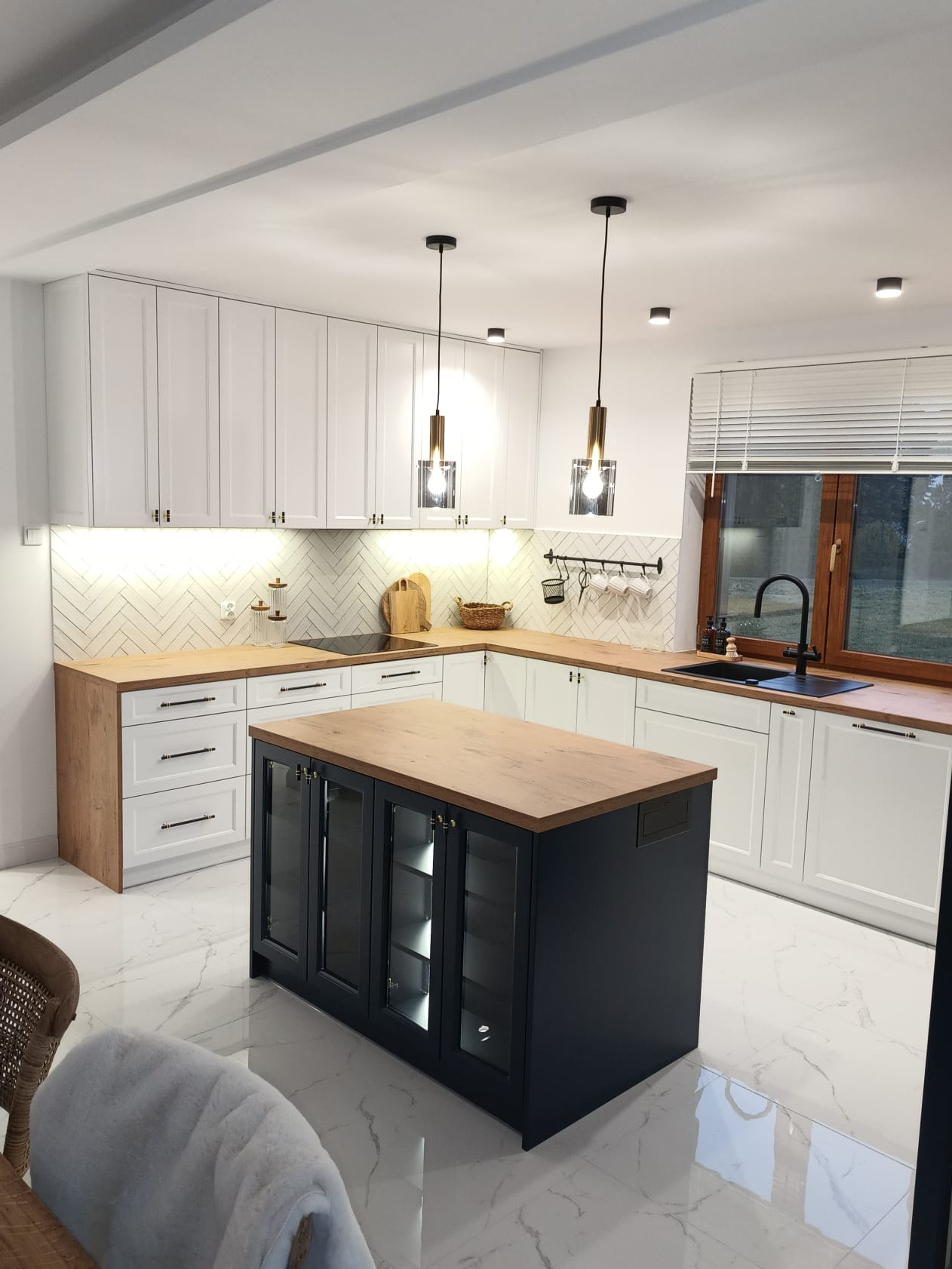 Nowoczesna kuchnia z białą zabudową, drewnianym blatem i czarną wyspą z przeszklonymi szafkami, oświetlona wiszącymi lampami z elementami mosiądzu.