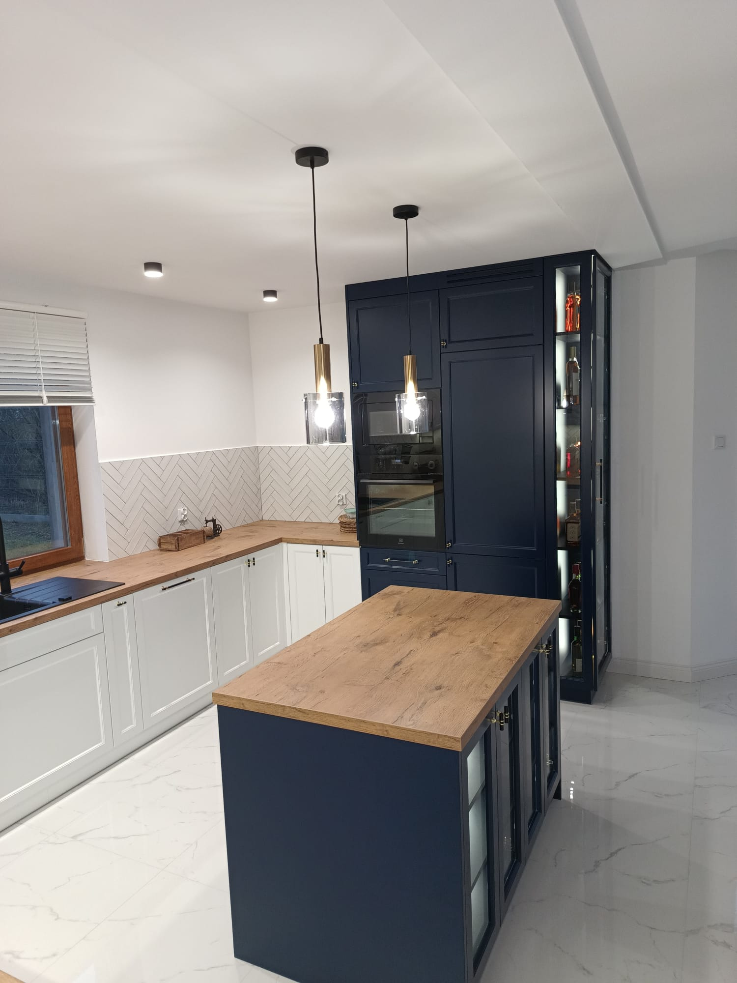 Nowoczesna kuchnia z biało-granatowymi szafkami, drewnianym blatem i wyspą kuchenną podświetloną wiszącymi lampami, na podłodze białe płytki imitujące marmur.