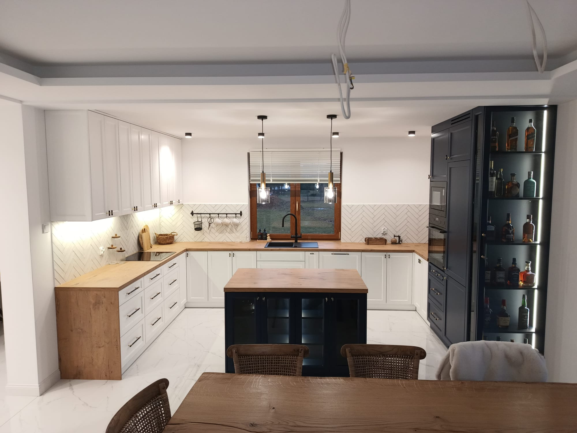 Przestronna kuchnia z białymi szafkami i drewnianymi blatami, wyspa kuchenna w kolorze granatowym z drewnianym blatem, podłoga z białego marmuru, na ścianie białe płytki ułożone w jodełkę, w tle...