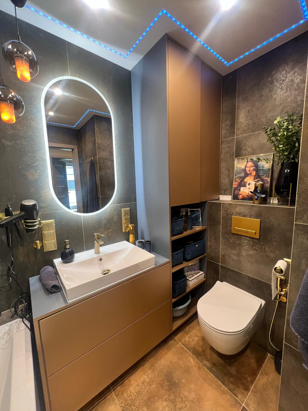 Nowoczesna łazienka z szarobrązowymi płytkami, podświetlanym lustrem i złotymi akcentami, w tym armatura i rama obrazu z Mona Lisą.