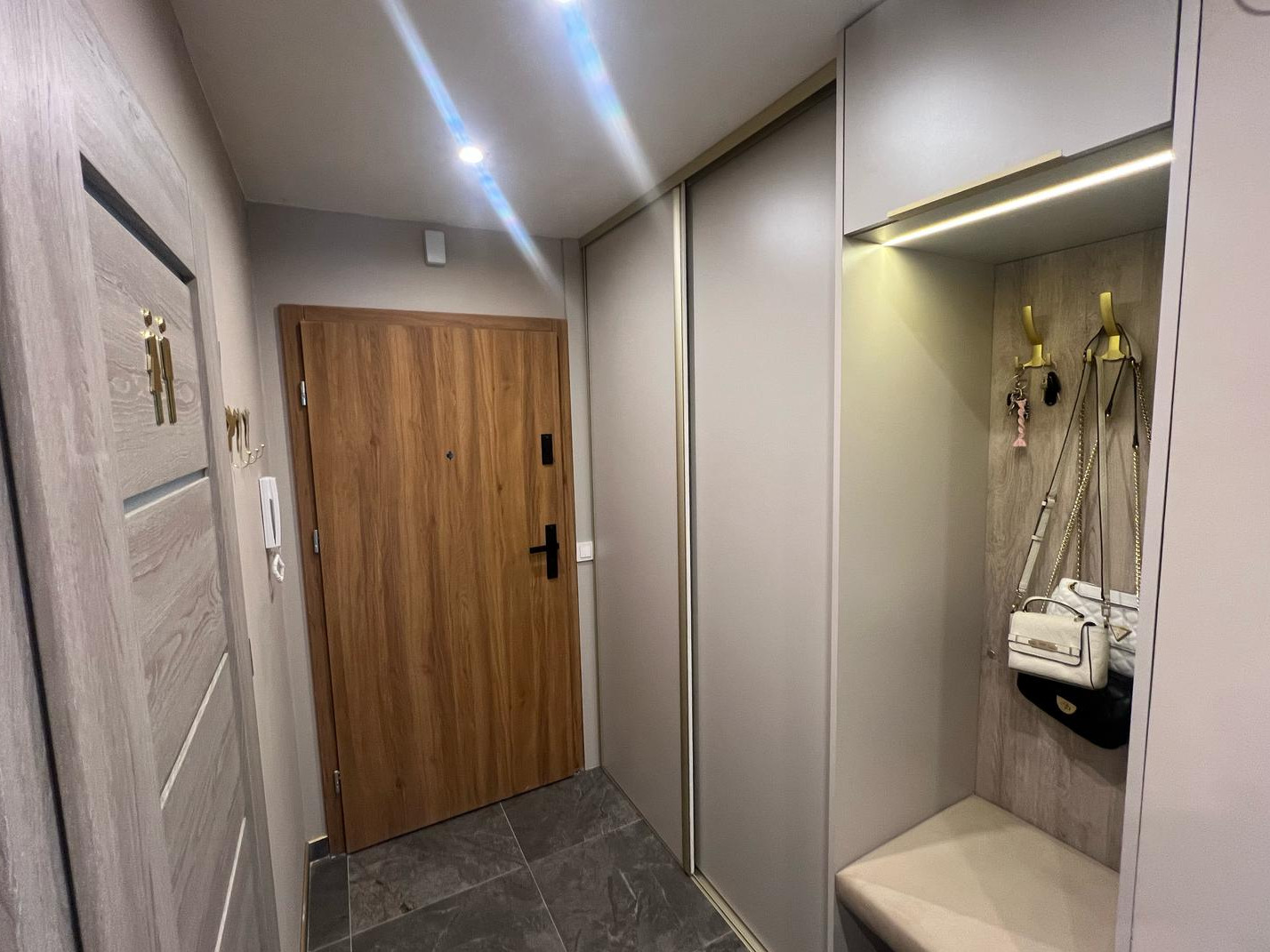 Przedpokój z przesuwną szafą wnękową w odcieniach szarości, oświetleniem LED i drewnianymi drzwiami wejściowymi, widoczne torebki i klucze na złotych haczykach.