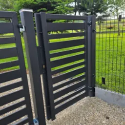 Nowoczesna, czarna furtka z poziomymi szczeblami, zamontowana obok ogrodzenia z siatki. Furtka otwarta, widoczny mechanizm zamka i podłoże z kamieni.