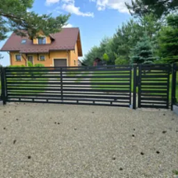 Nowoczesna brama wjazdowa, czarna, poziome profile, na tle domu z brązowym dachem i zieleni. Brama zamontowana na żwirowym podłożu, z prawej strony furtka.