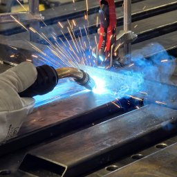 BACHURSKI SPÓŁKA Z OGRANICZONĄ ODPOWIEDZIALNOŚCIĄ - Spawanie metalowych profili na perforowanym stole spawalniczym; widoczne iskry, dym i rękawica ochronna. Czerwony uchwyt trzyma element w miejscu.