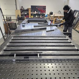 BACHURSKI SPÓŁKA Z OGRANICZONĄ ODPOWIEDZIALNOŚCIĄ - Spawanie metalowej bramy na perforowanym stole spawalniczym w warsztacie. Widoczny spawacz w trakcie pracy, obok elementy ramy i narzędzia.