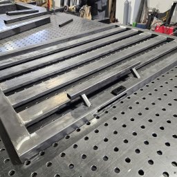 BACHURSKI SPÓŁKA Z OGRANICZONĄ ODPOWIEDZIALNOŚCIĄ - Metalowa rama bramy w trakcie spawania na perforowanym stole spawalniczym. Widoczne narzędzia i elementy konstrukcyjne w warsztacie.