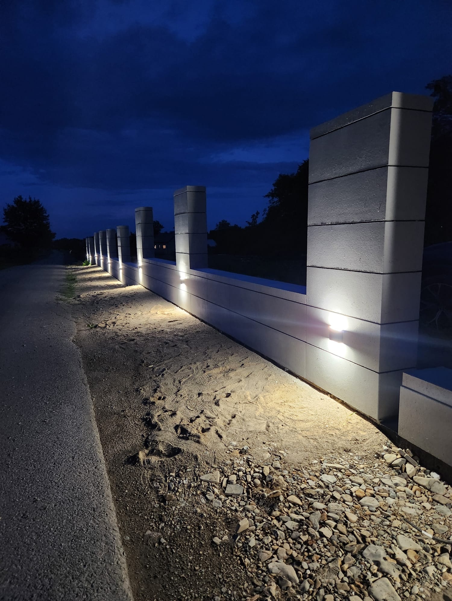 Podświetlony fragment długiego muru z betonowych bloczków, z wbudowanymi lampkami, widziany nocą z perspektywy gruntu.