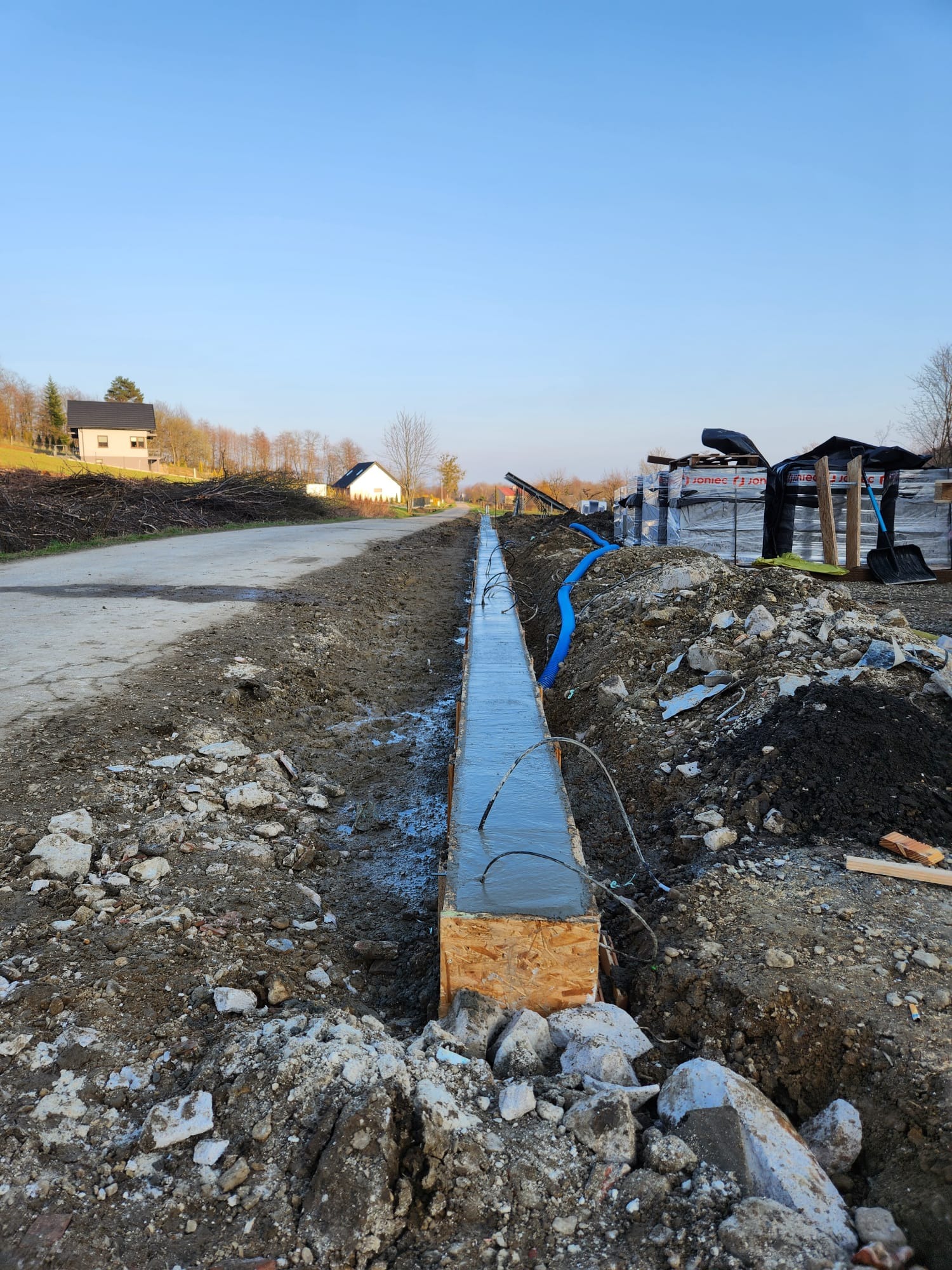 Wykopany rów z drewnianym szalunkiem wypełniony świeżym betonem, widoczne przewody i niebieska rura, w tle ułożone palety z materiałami budowlanymi i domy.