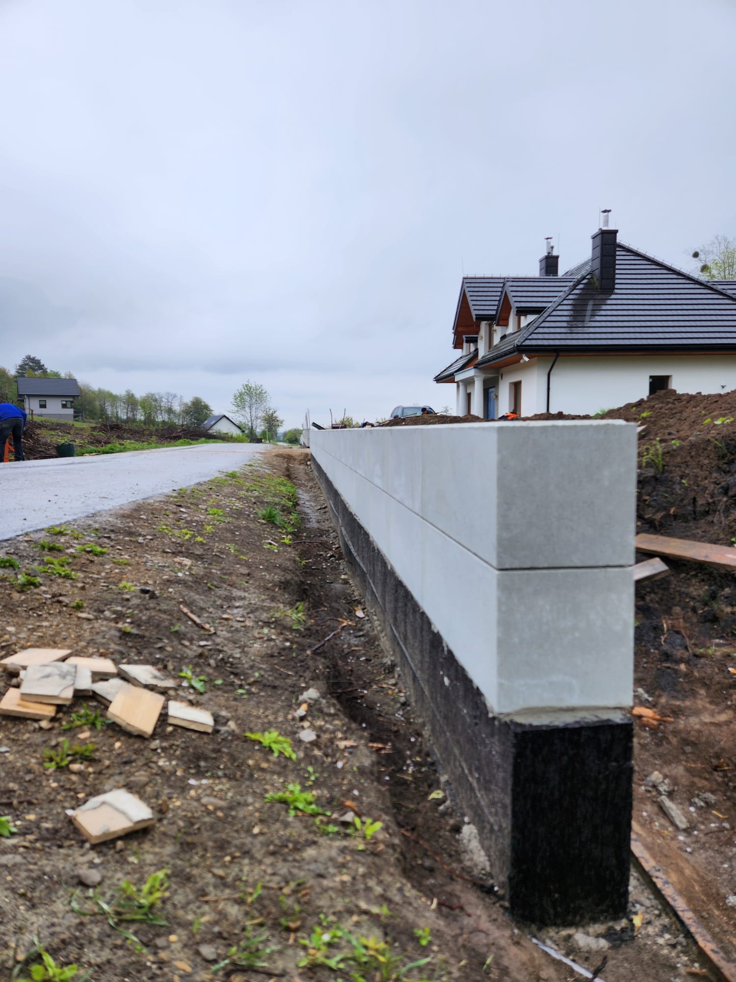 Wznoszenie murku oporowego z prefabrykatów betonowych, widoczne hydroizolacja fundamentu, fragment drogi i budynek w tle, pochmurny dzień.