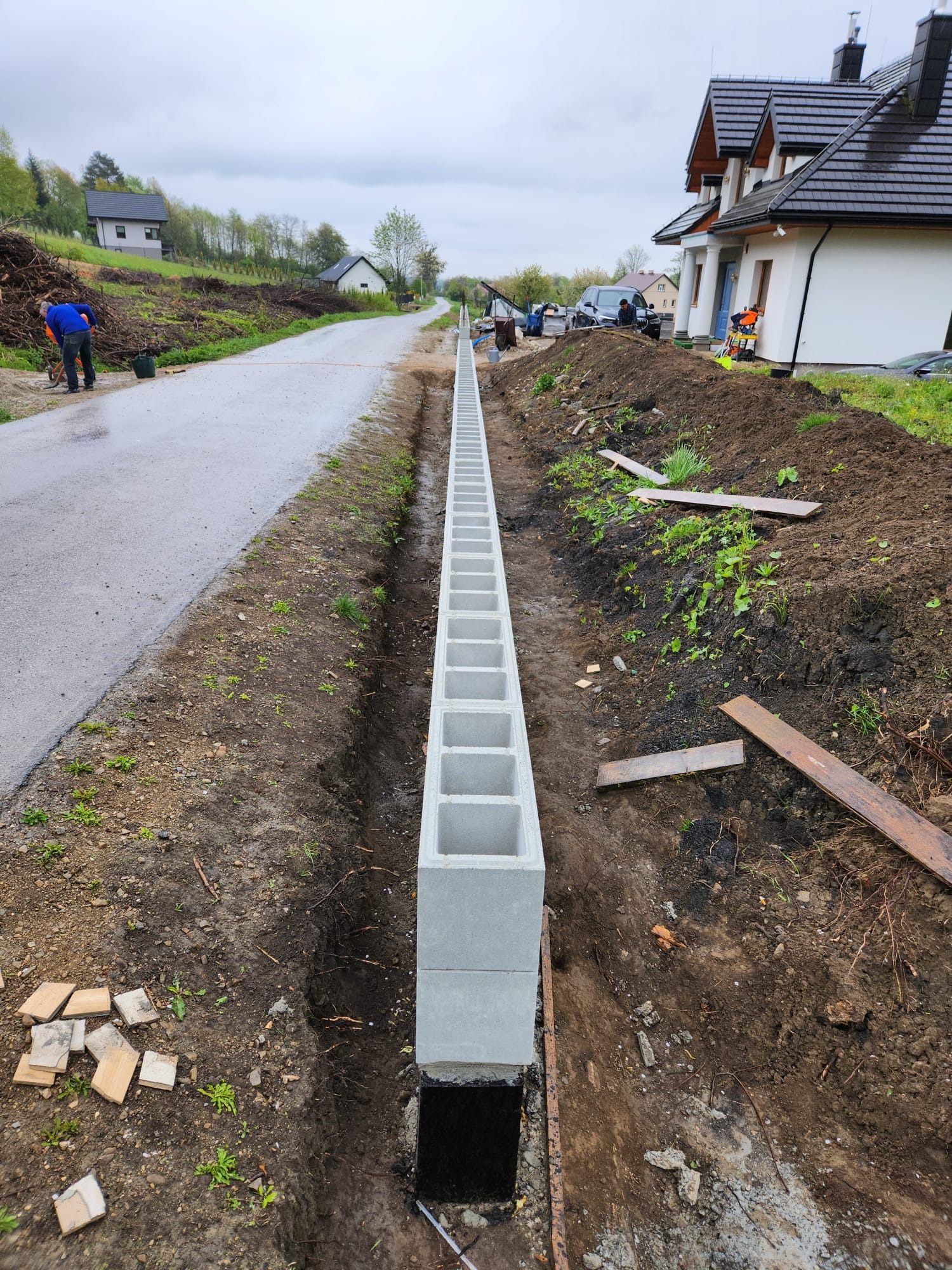 Wąski fundament pod ogrodzenie z szarych, betonowych bloczków, widok z góry na wykop w ziemi, obok asfaltowa droga i dom w tle.
