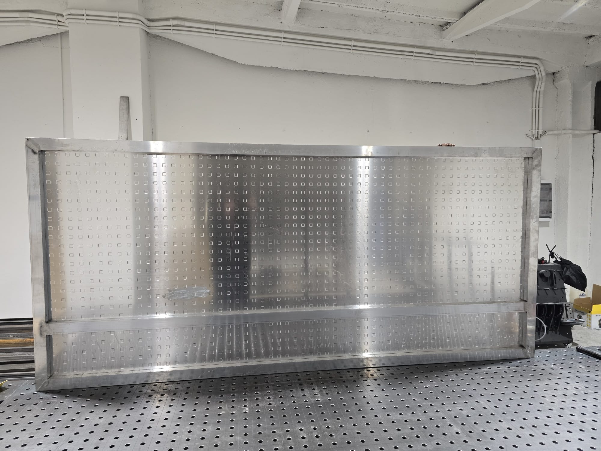 Metalowa konstrukcja bramy wjazdowej z perforacją kwadratową, leżąca na perforowanym stole warsztatowym w jasnym pomieszczeniu. Widoczne spawane łączenia ramy.