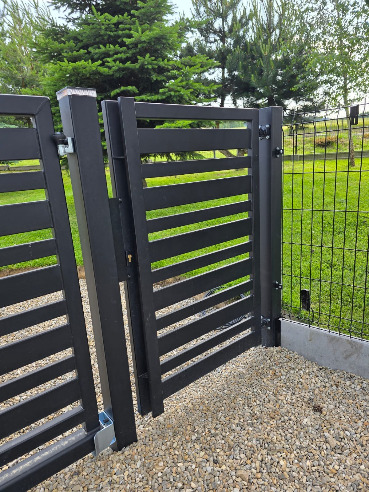 Nowoczesna, czarna furtka z poziomymi szczeblami, zamontowana obok ogrodzenia z siatki. Furtka otwarta, widoczny mechanizm zamka i podłoże z kamieni.