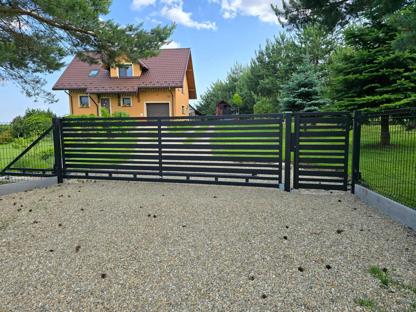 Nowoczesna brama wjazdowa, czarna, poziome profile, na tle domu z brązowym dachem i zieleni. Brama zamontowana na żwirowym podłożu, z prawej strony furtka.