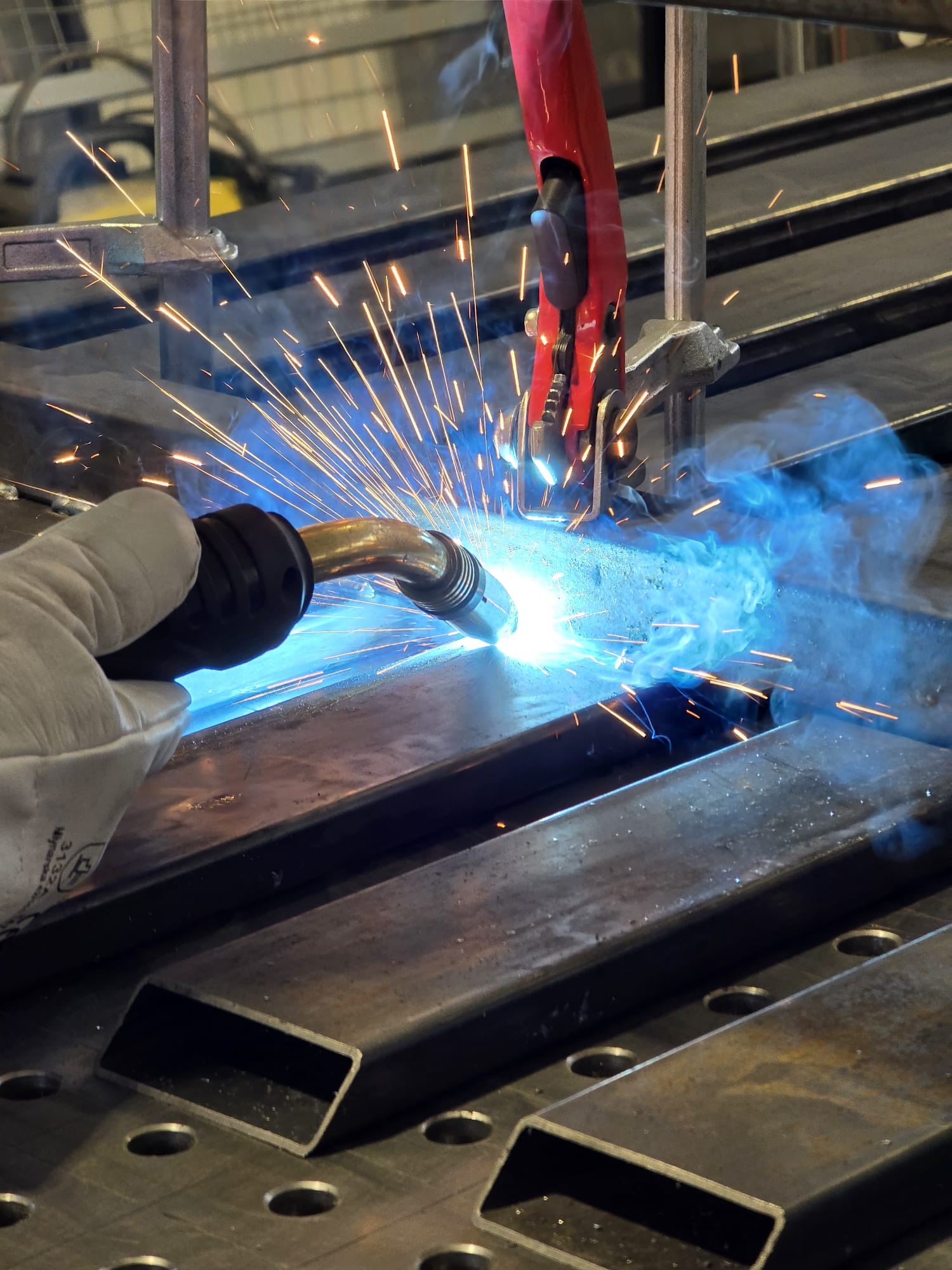 Spawanie metalowych profili na perforowanym stole spawalniczym; widoczne iskry, dym i rękawica ochronna. Czerwony uchwyt trzyma element w miejscu.
