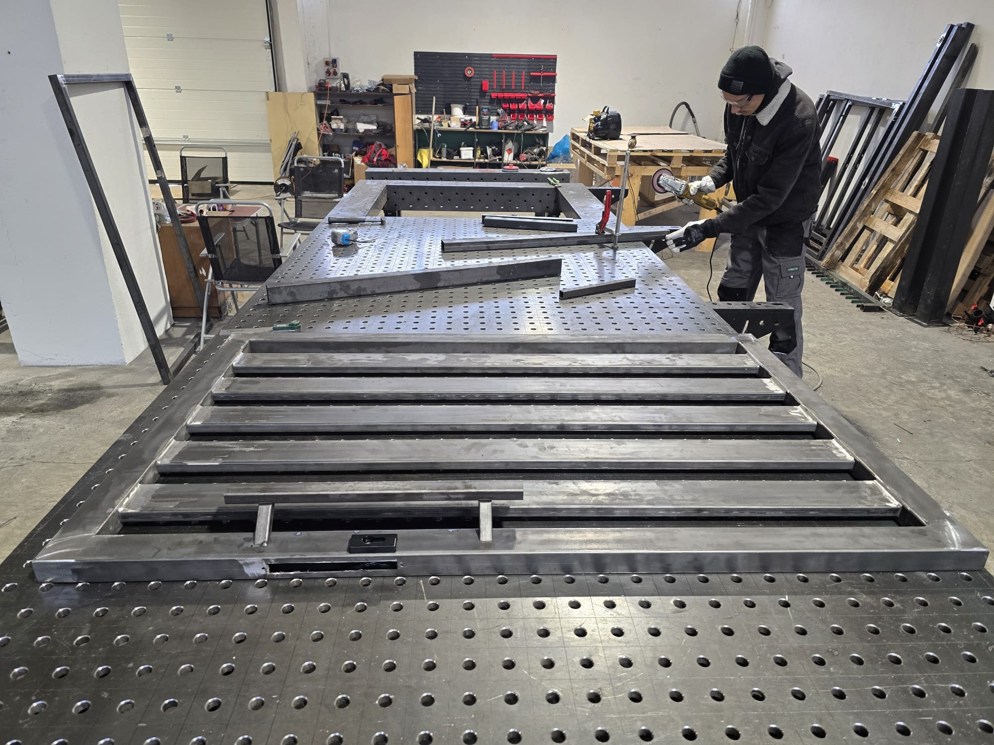 Spawanie metalowej bramy na perforowanym stole spawalniczym w warsztacie. Widoczny spawacz w trakcie pracy, obok elementy ramy i narzędzia.