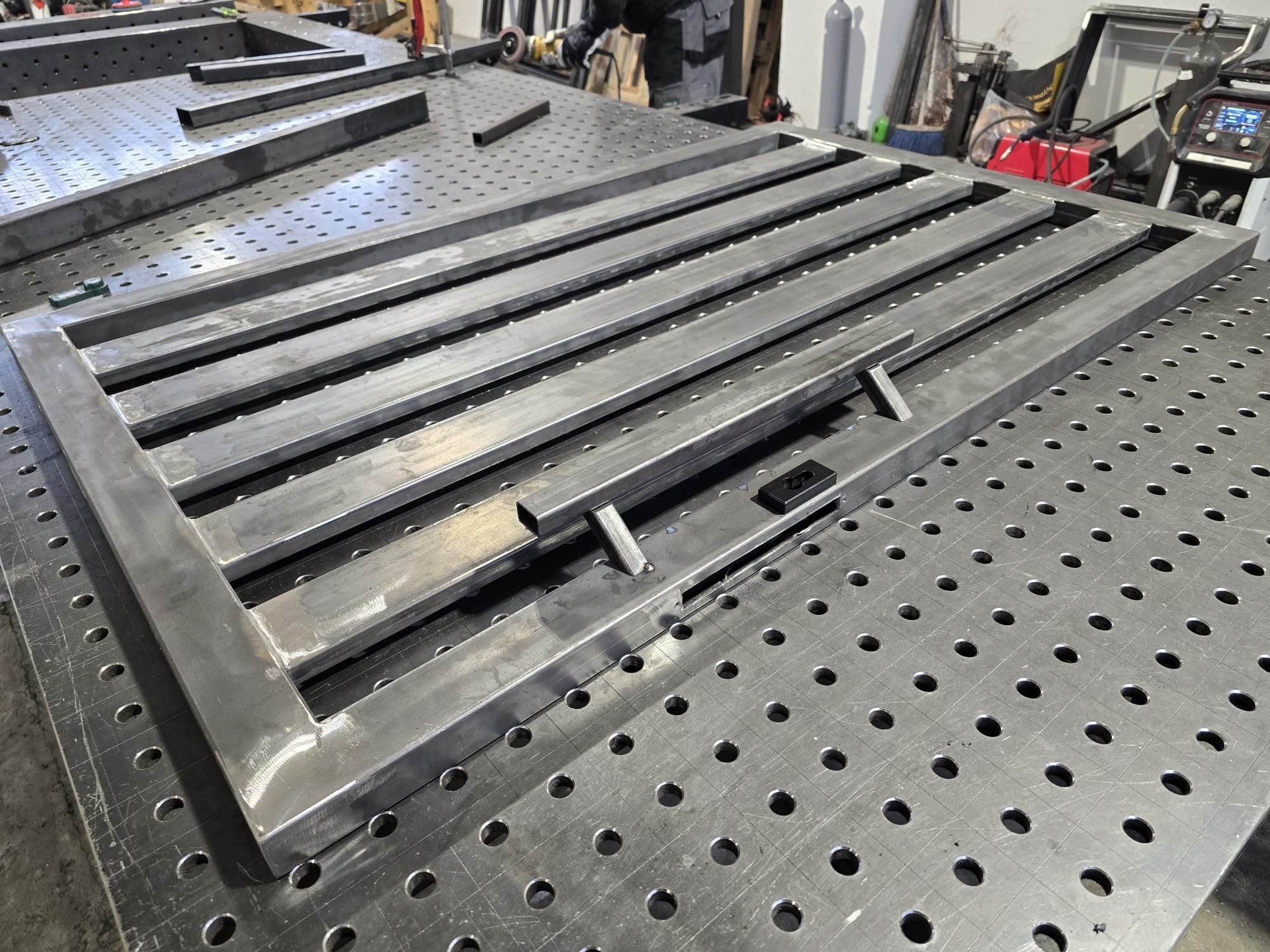 Metalowa rama bramy w trakcie spawania na perforowanym stole spawalniczym. Widoczne narzędzia i elementy konstrukcyjne w warsztacie.