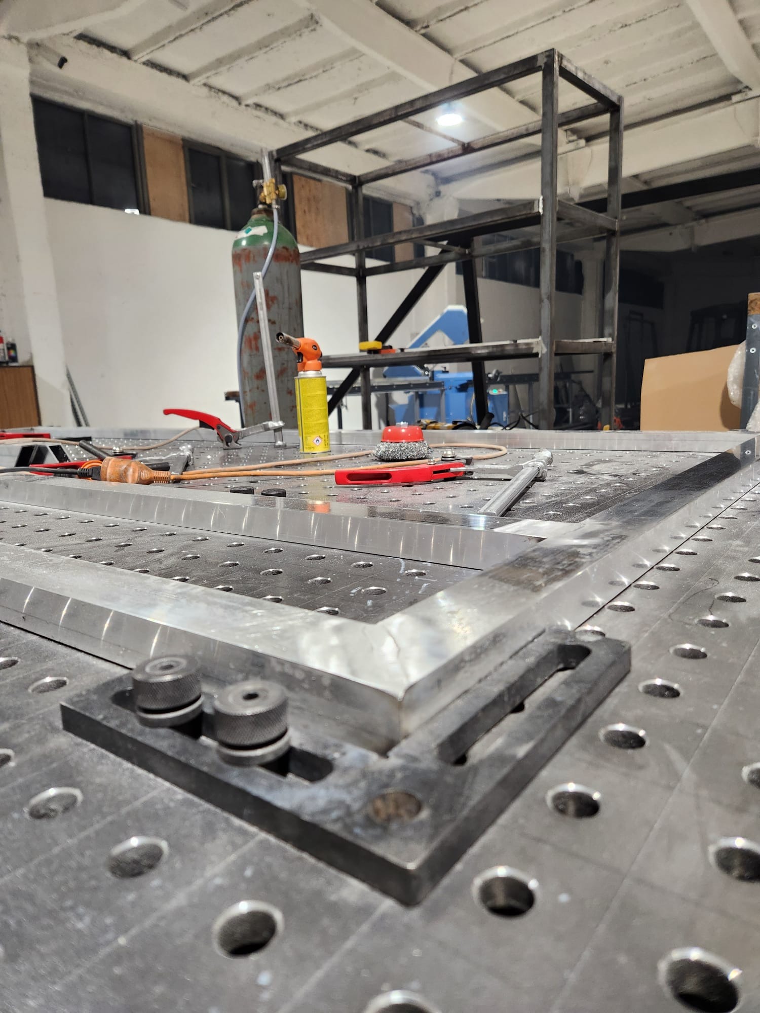 Metalowa konstrukcja w trakcie spawania na perforowanym stole spawalniczym. W tle butla z gazem i inne narzędzia.