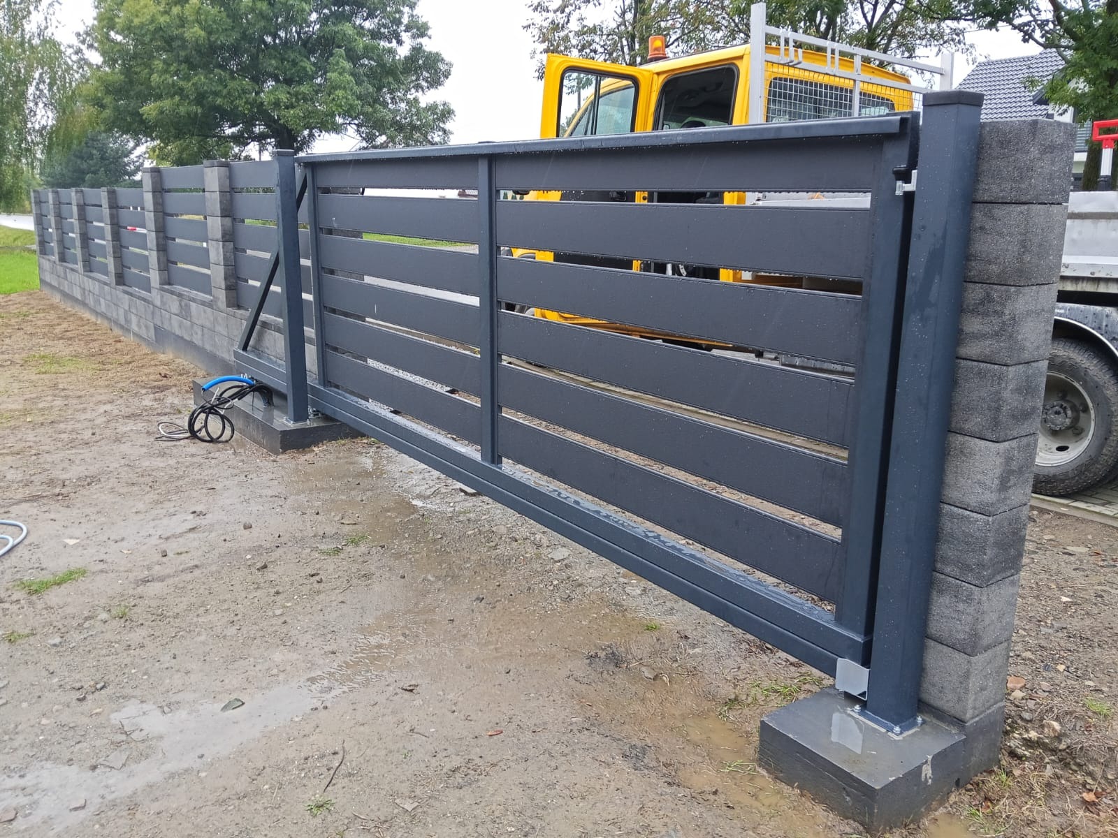 Nowoczesna, przesuwna brama wjazdowa z poziomymi panelami, osadzona na betonowych słupkach. Widoczne elementy montażowe i fragment pojazdu w tle.