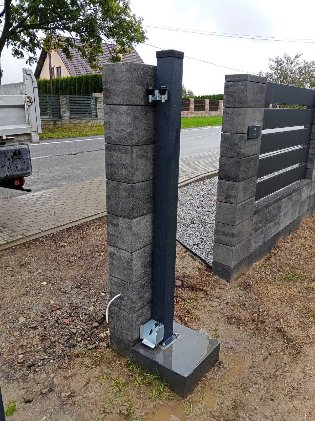 Szary, betonowy słupek bramy przesuwnej z zamontowanym mechanizmem, widoczny fragment ogrodzenia panelowego i podłoże z drobnym kamieniem.