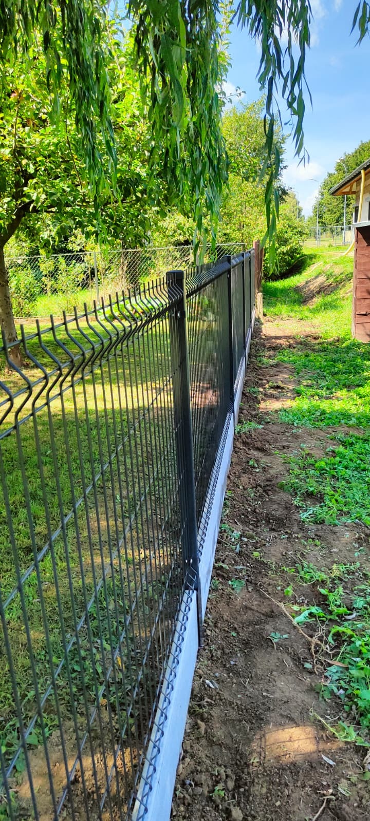 Nowoczesne ogrodzenie panelowe w kolorze grafitowym, osadzone na niskiej, betonowej podmurówce. Widok na posesję z zielonym trawnikiem i drzewami w tle.