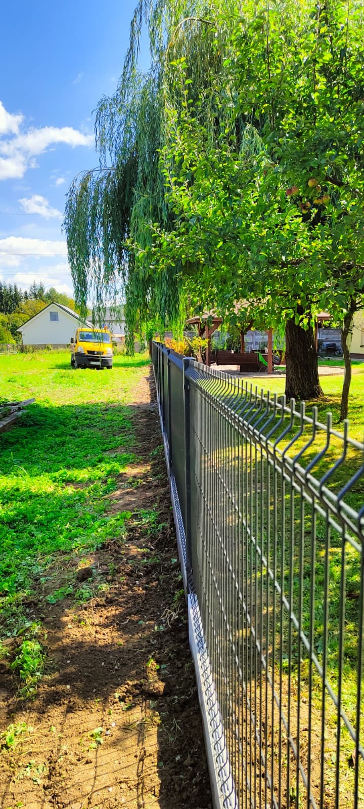 Nowo postawione ogrodzenie panelowe wzdłuż działki, z widocznym betonowym podmurówką. W tle zielona trawa, drzewa i budynek. Żółty samochód dostawczy parkuje w oddali.