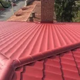 Czerwony dach z blachy trapezowej z widocznym kominem z cegły i rynną, na tle zabudowy miejskiej.