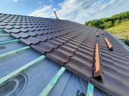 Nowy, brązowy dach z blachodachówki ułożony na drewnianej konstrukcji, widok z góry, w tle zielone wzgórze i błękitne niebo z chmurami.