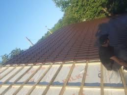 Układanie brązowej dachówki ceramicznej na przygotowanej konstrukcji drewnianej z widoczną izolacją, w tle zieleń drzew i błękitne niebo, ujęcie z dołu ukazujące postęp prac.