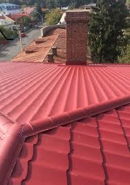 Czerwony dach z blachy trapezowej z widocznym kominem z cegły i rynną, na tle zabudowy miejskiej.