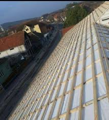 Konstrukcja dachu z drewnianych łat i kontrłat pokryta białą membraną dachową, widok z góry na tle zabudowań mieszkalnych.